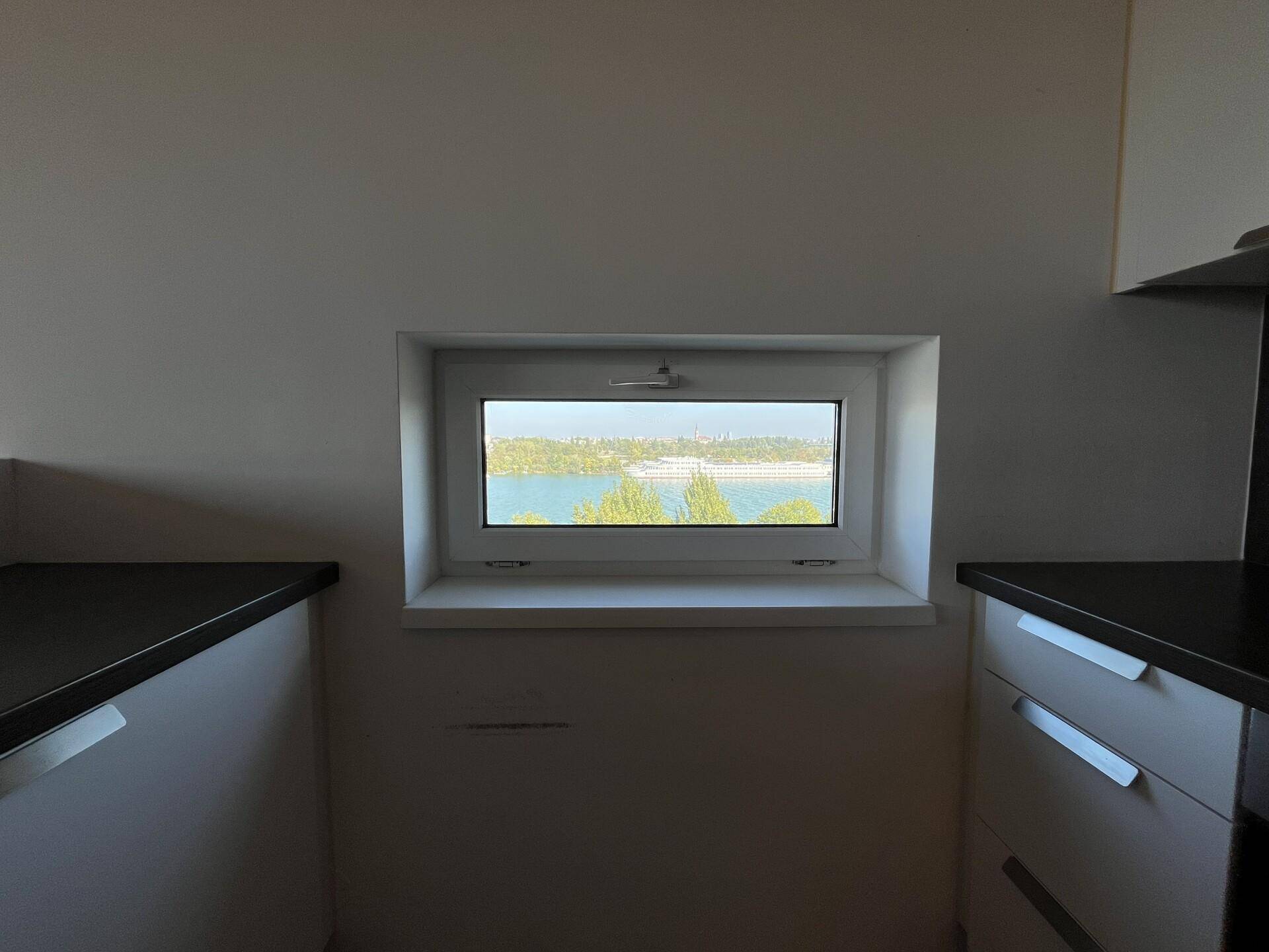 Kleines Küchenfenster mit Ausblick auf das Wasser und grüne Bäume, umgeben von dunklen Arbeitsplatten.