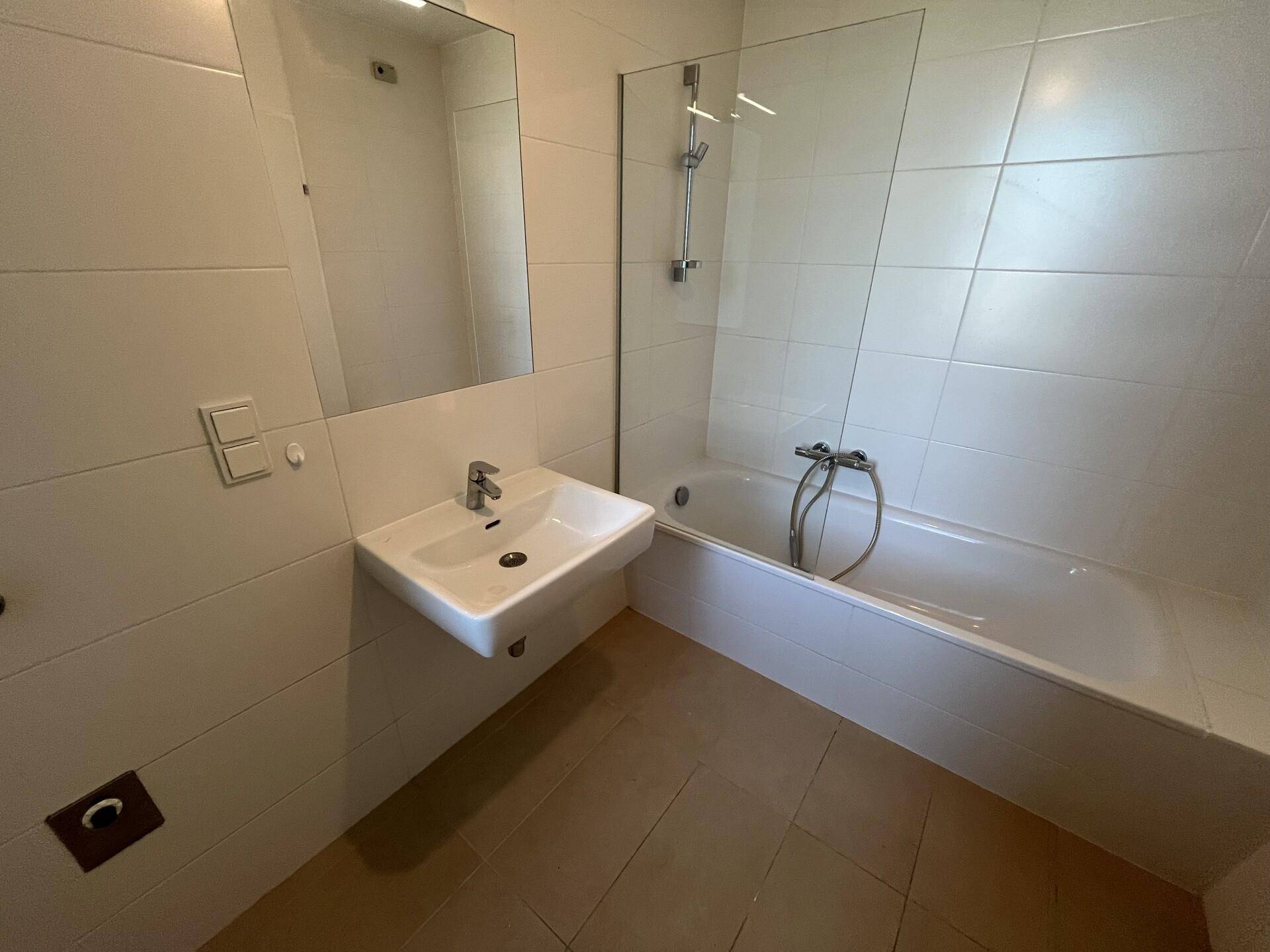 Helles Badezimmer mit weißen Fliesen, Badewanne mit Duschwand und modernem Waschbecken.