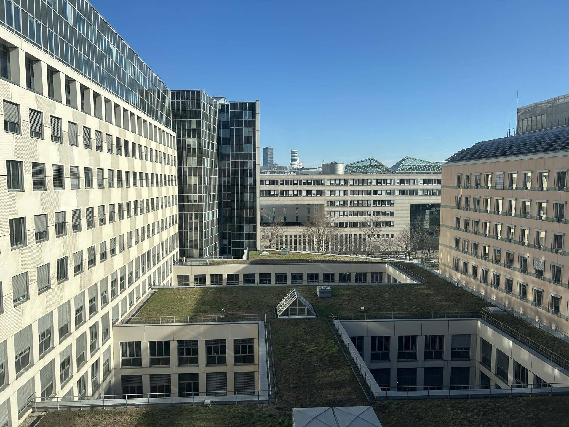 Ausblick von einem oberen Stockwerk auf moderne Gebäude mit begrünten Dächern und die Stadt im Hintergrund.
