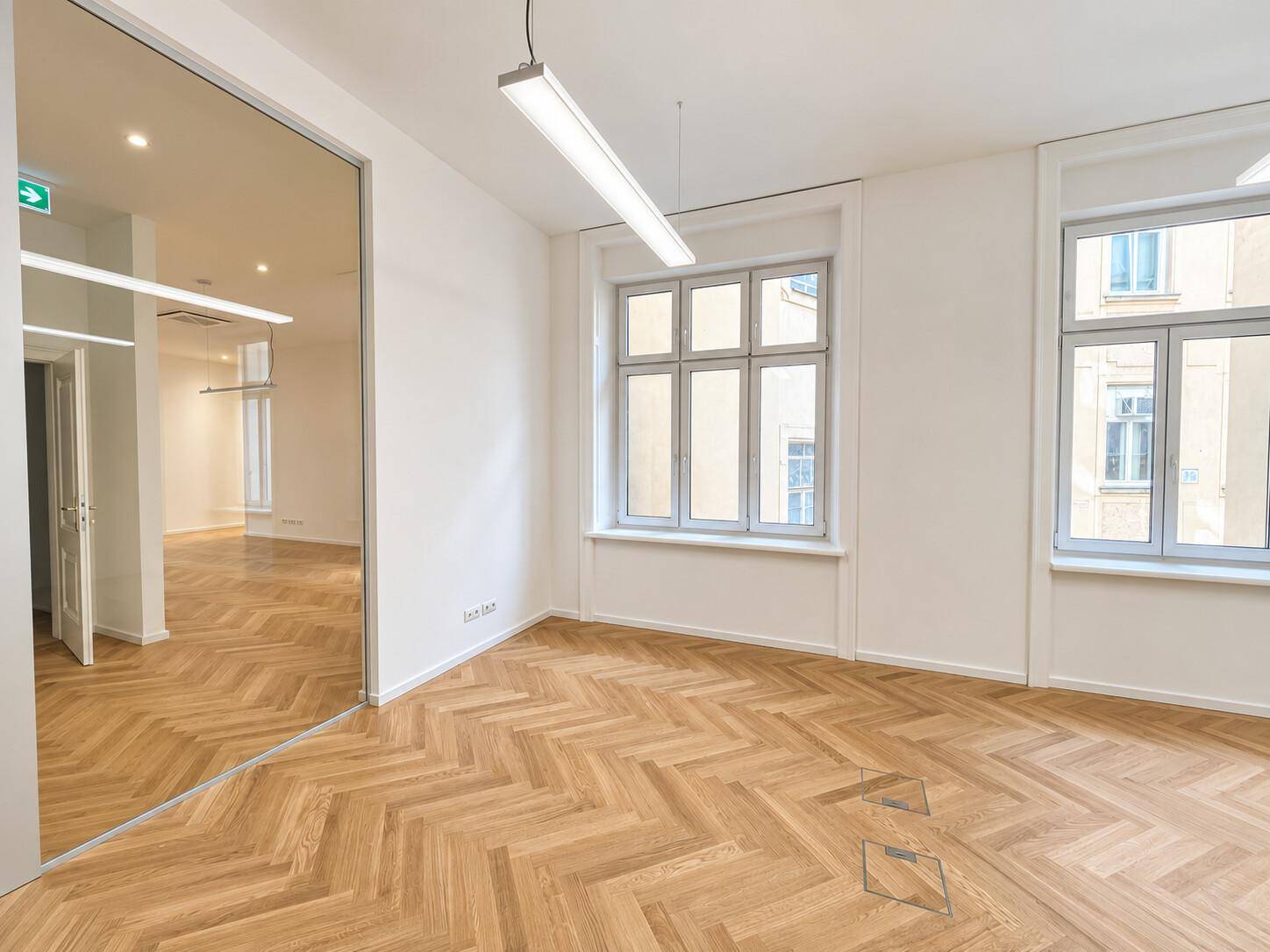 Heller Büroraum mit Fischgrätparkett, großen Fenstern und einer verspiegelten Wand.