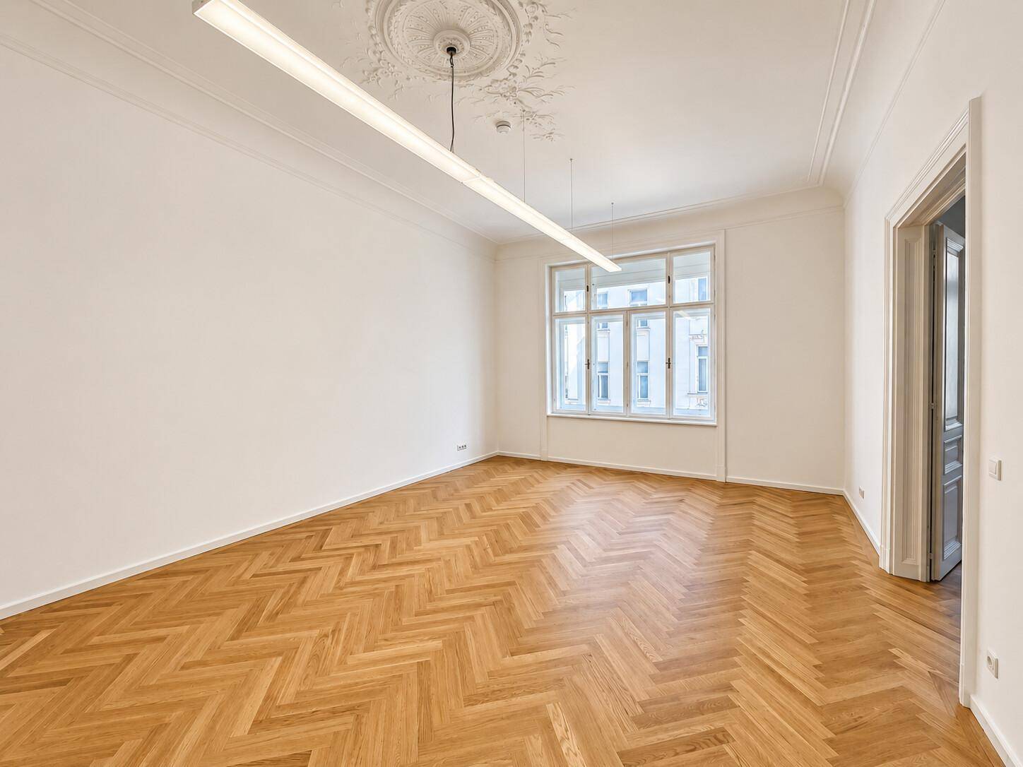 Leerer Raum mit klassischem Fischgrätparkett, hohen Decken und einem großen Fenster.