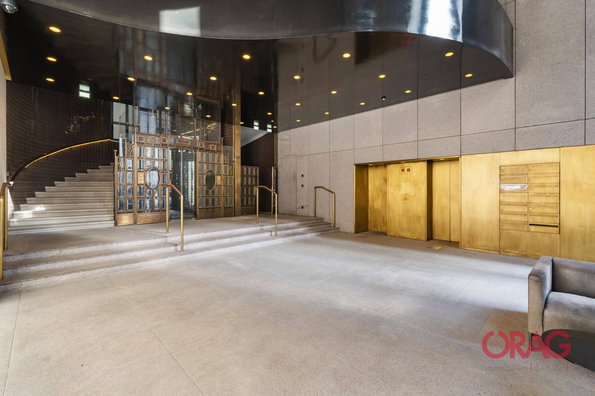 Elegante Lobby mit goldfarbenen Aufzügen, einer breiten Steintreppe und einem markanten, verzierten Eingangsbereich.