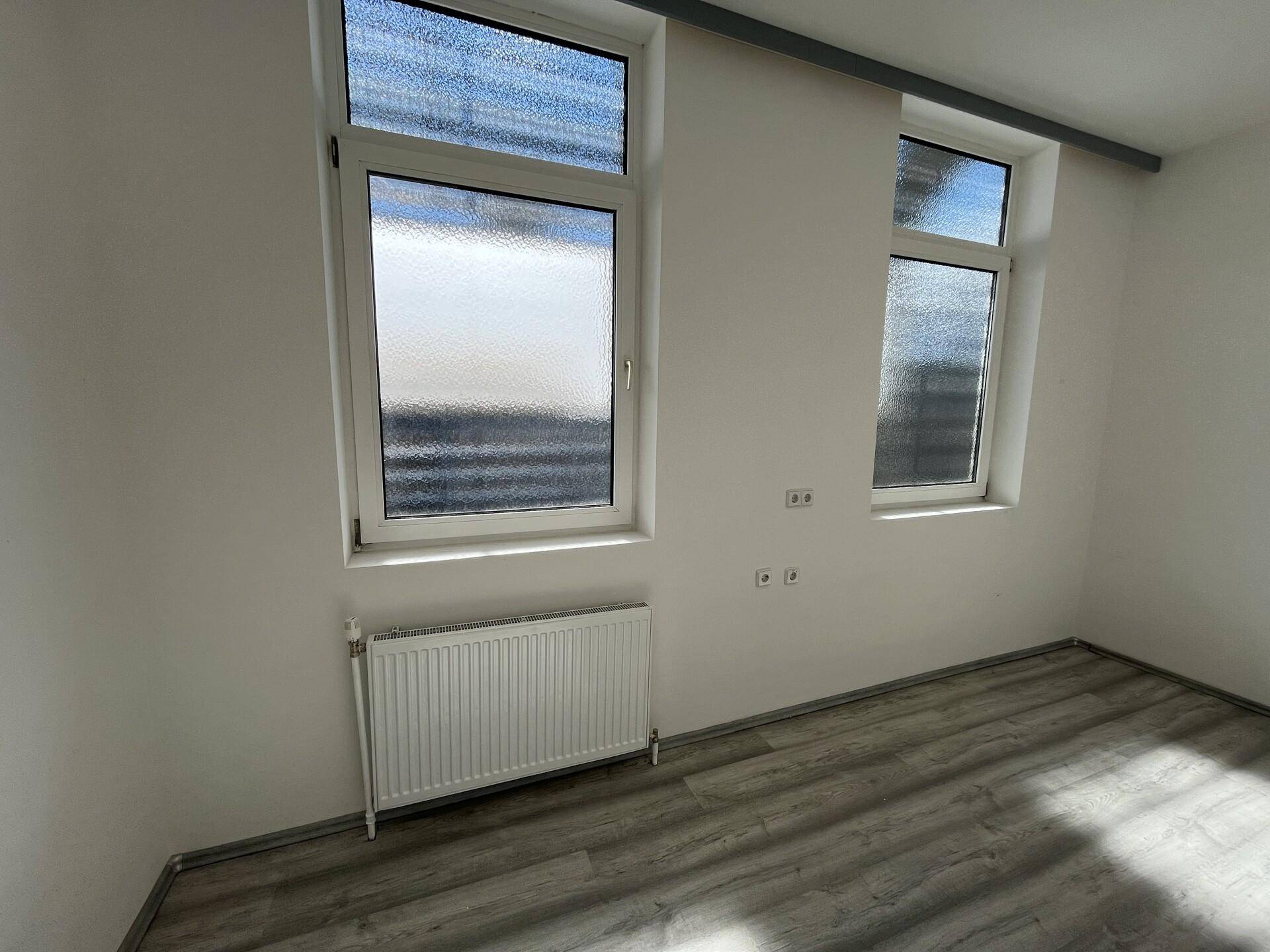 Kleinerer Raum mit zwei Fenstern, Heizkörper und hellem Laminatboden, ideal als Büro oder Lager.