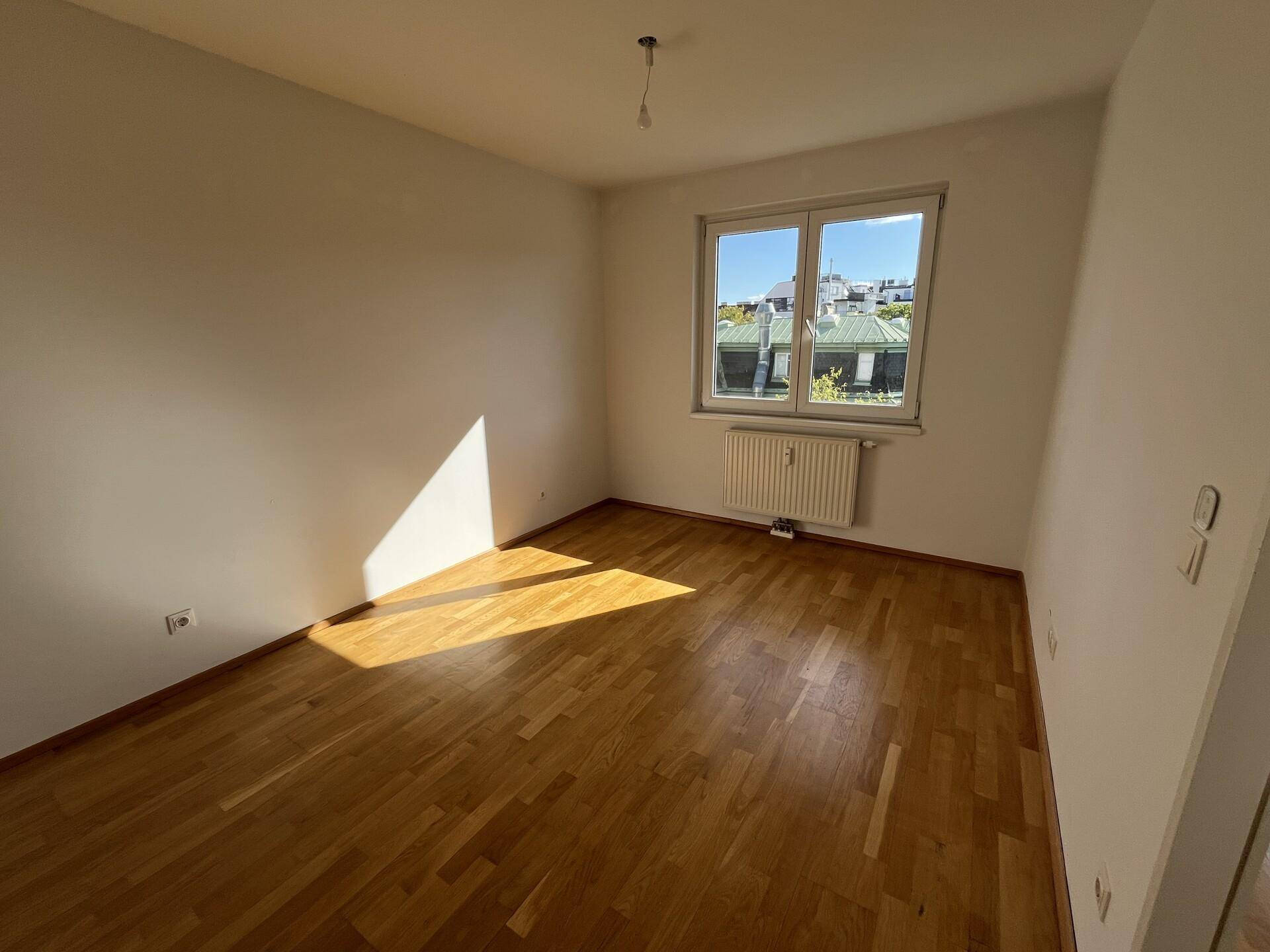 Helles Schlafzimmer mit Parkettboden und einem Fenster, das viel Tageslicht hereinlässt.