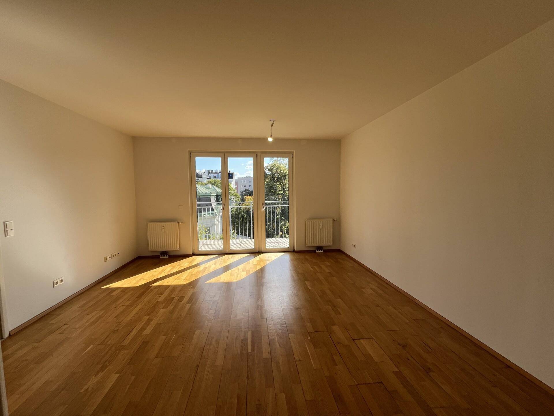 Geräumiges Wohnzimmer mit Parkettboden und Zugang zu einem Balkon, durchflutet von Sonnenlicht.