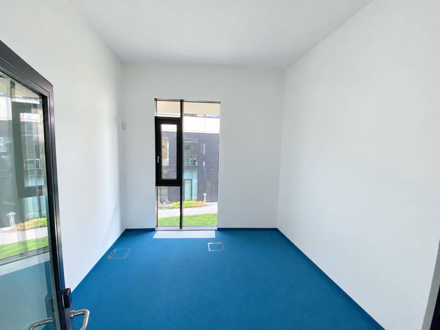 Kleiner, heller Büroraum mit blauem Teppichboden und einer großen Fensterfront zum Außenbereich.