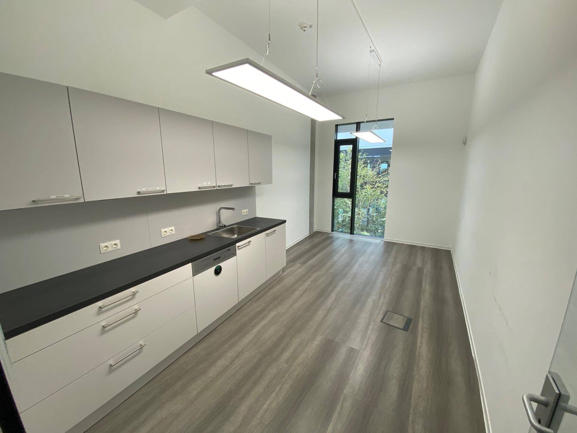 Moderne Teeküche mit hellgrauen Schränken, dunkler Arbeitsplatte und Vinylboden in Holzoptik.