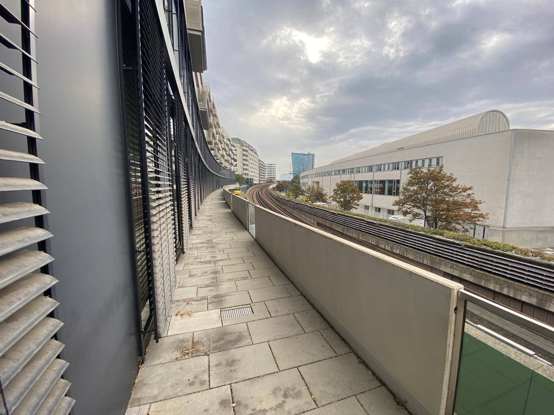 Langer, schmaler Außenbereich mit Steinplattenboden und Blick auf die Bahngleise und Stadt.