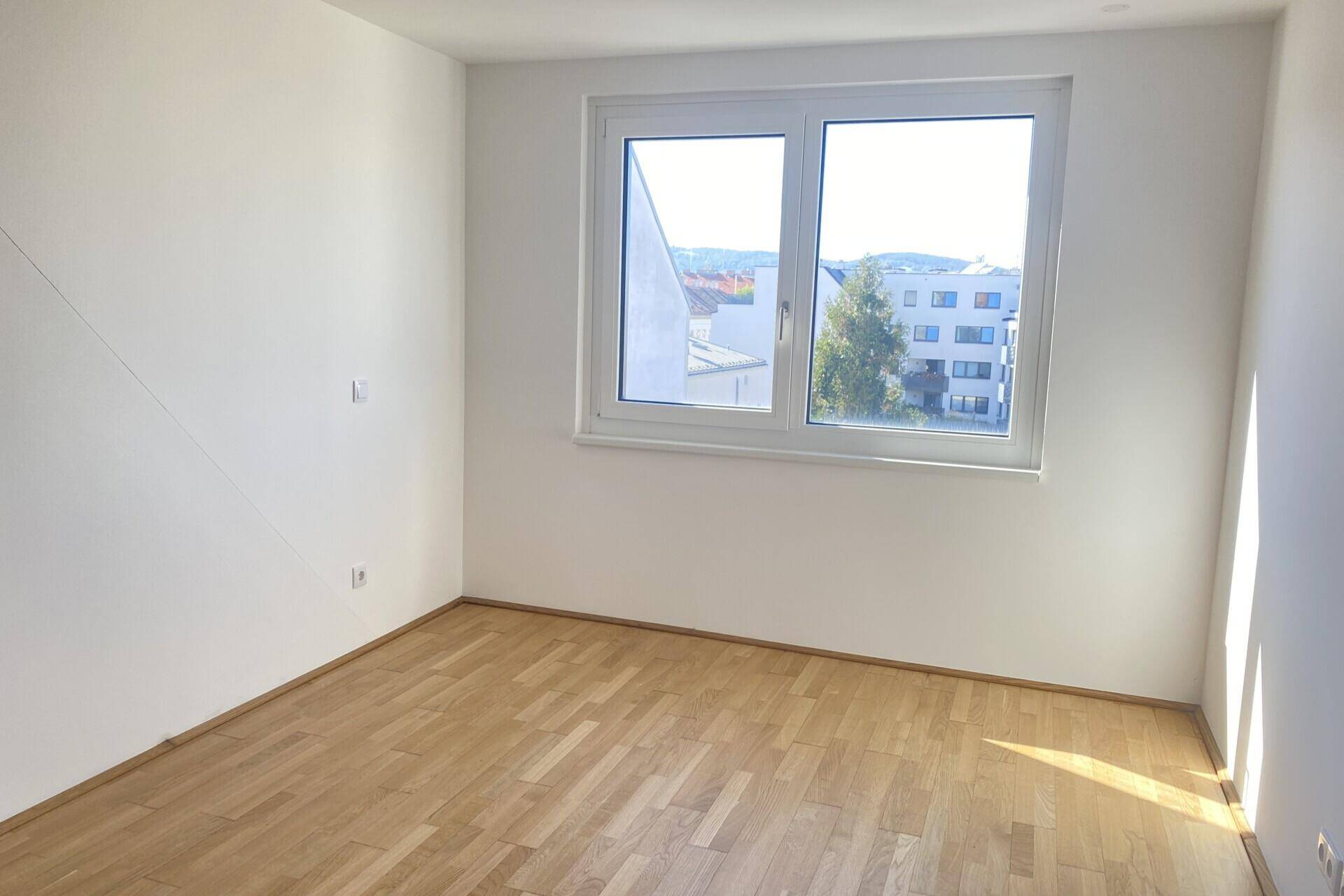 Helles Schlafzimmer mit Parkettboden und großem Fenster, das viel Tageslicht hereinlässt.