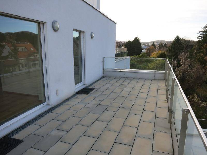 Geräumige Terrasse mit Fliesenboden und moderner Glasbrüstung, umgeben von Bäumen.