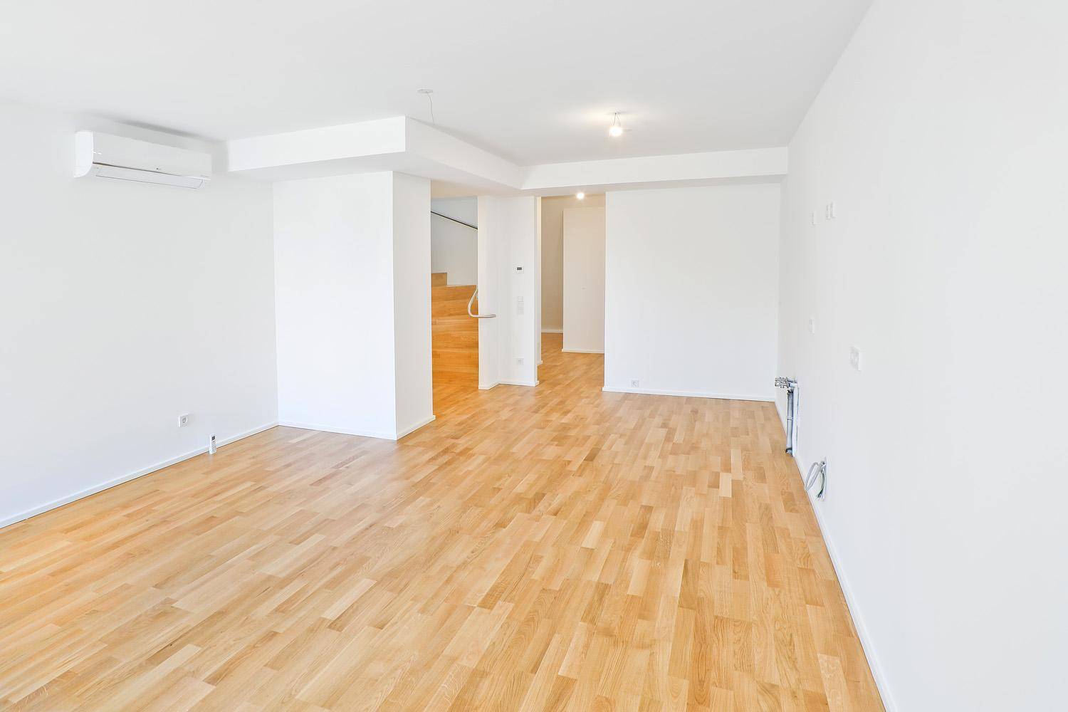 Geräumiger Wohnbereich mit hellem Parkettboden, weißen Wänden und Zugang zu einer Treppe.