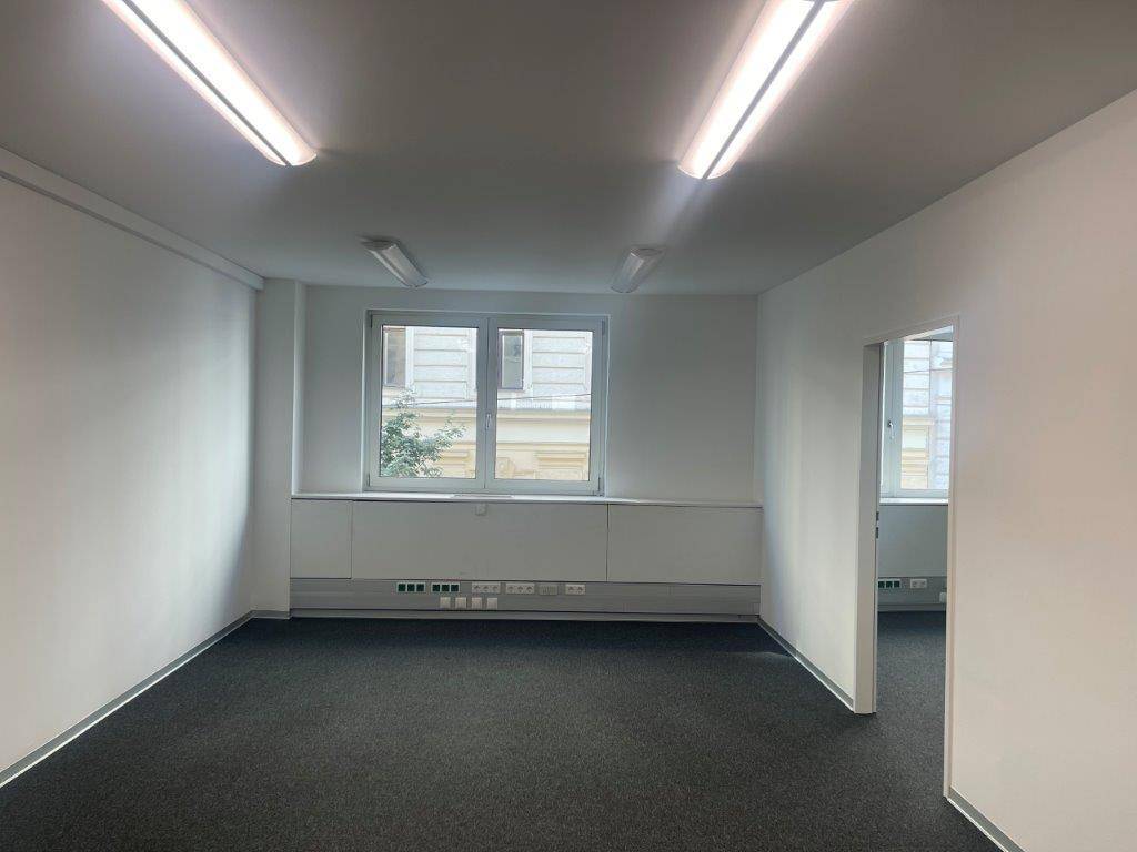 Leeres Büro mit Teppichboden, großen Fenstern und einem Durchgang zu einem angrenzenden Bereich.