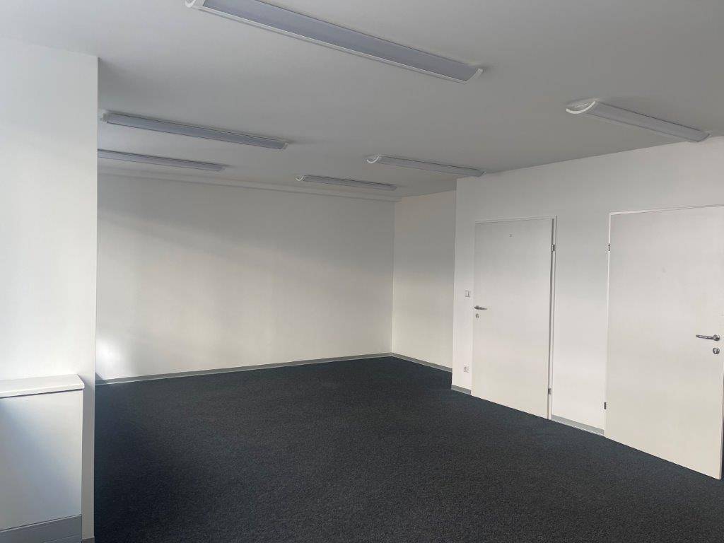 Großes, leeres Büro mit dunklem Teppichboden, weißen Wänden und zwei Türen.