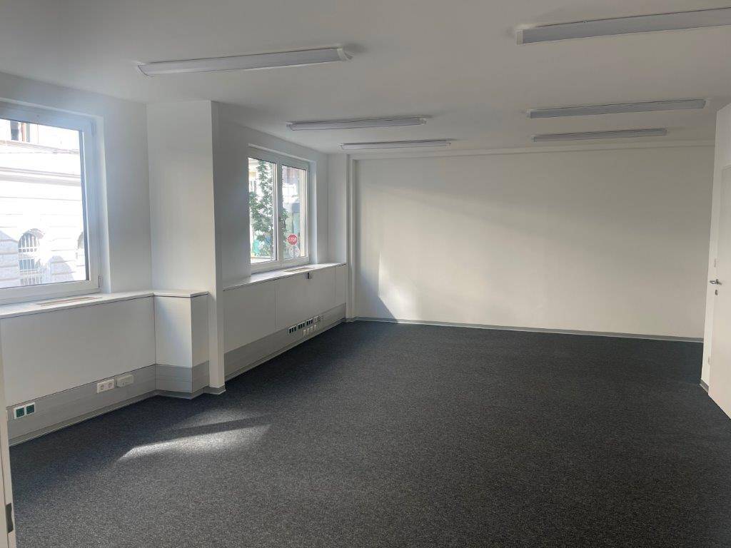 Geräumiges Büro mit Teppichboden, weißen Wänden und Fenstern, die viel Tageslicht spenden.