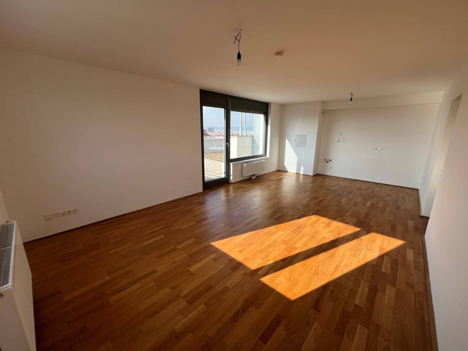 Helles, leeres Wohnzimmer mit Parkettboden und Zugang zu einem Balkon oder einer Terrasse.