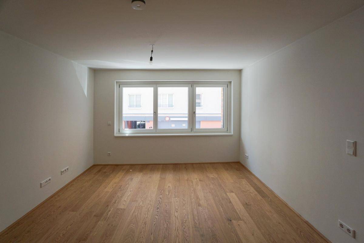 Geräumiges, leeres Wohnzimmer mit großen Fenstern, die viel Tageslicht hereinlassen, und hellem Holzfußboden.