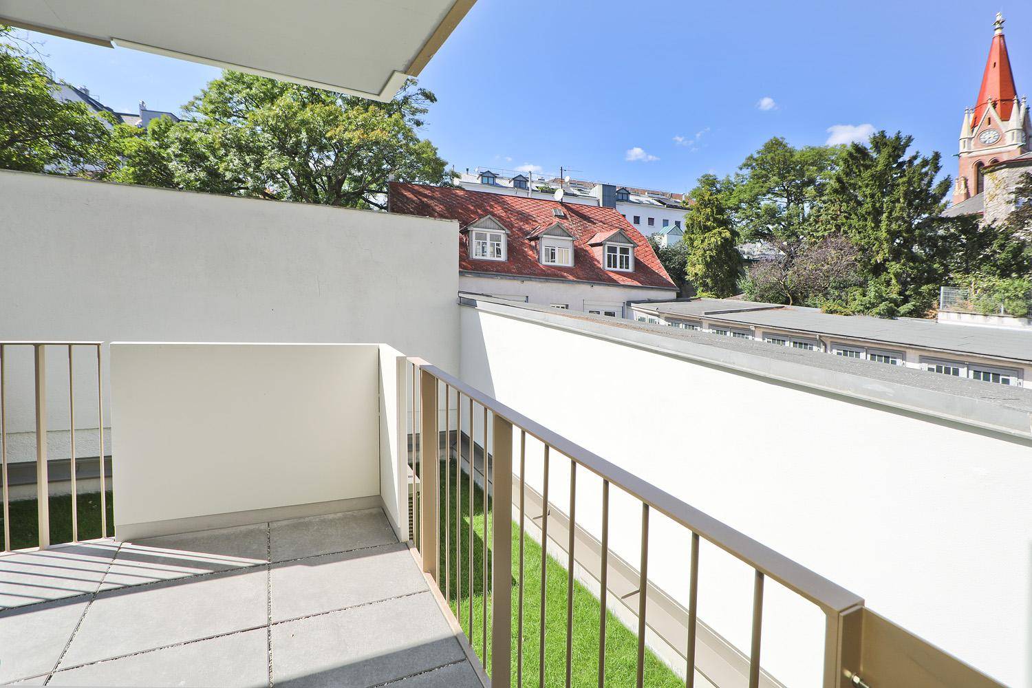 Sonniger Balkon mit Metallgeländer und Blick auf einen Kirchturm und grüne Bäume.