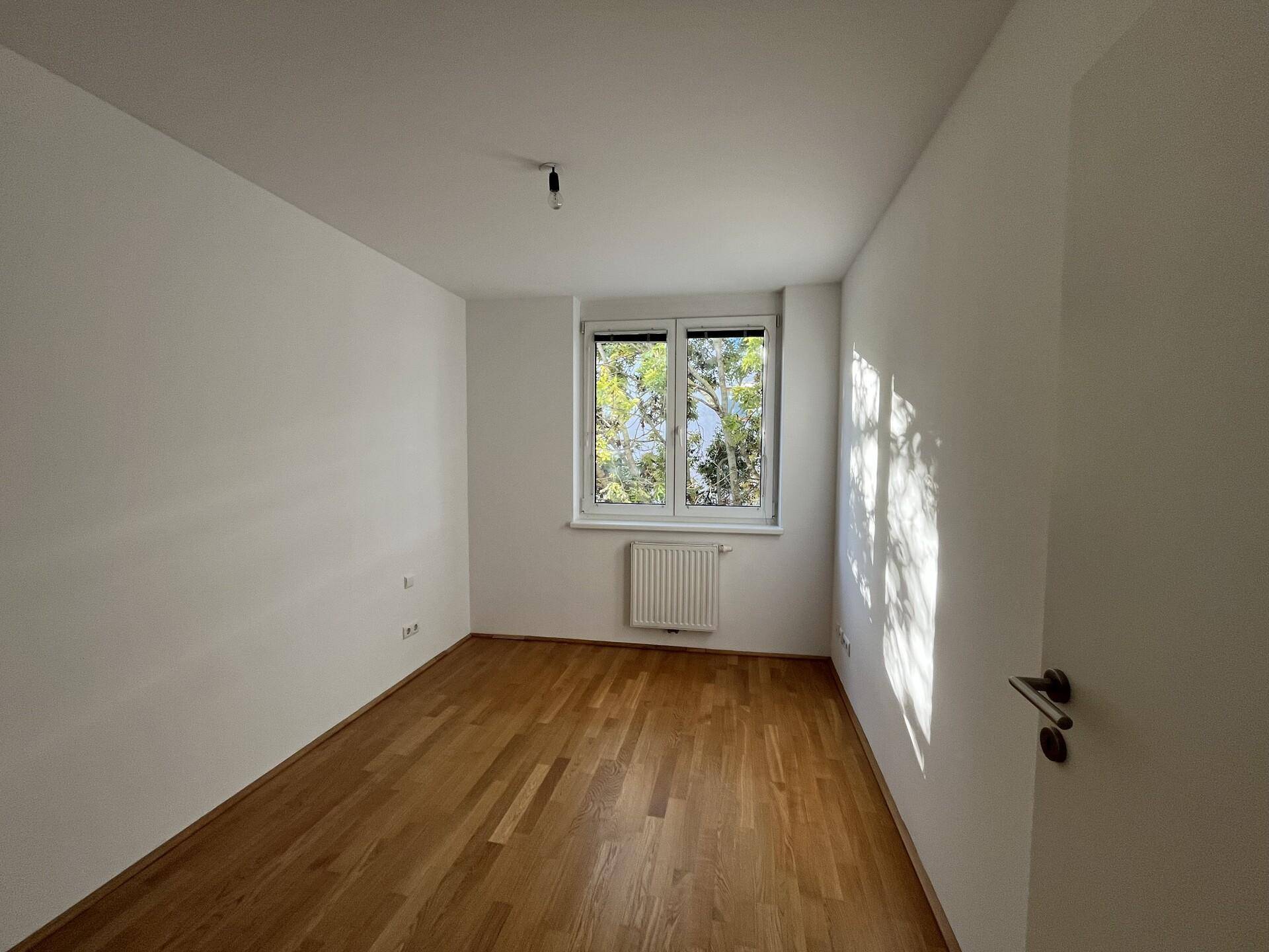 Geräumiges Zimmer mit hellem Parkettboden und einem Fenster, das natürliches Licht spendet.