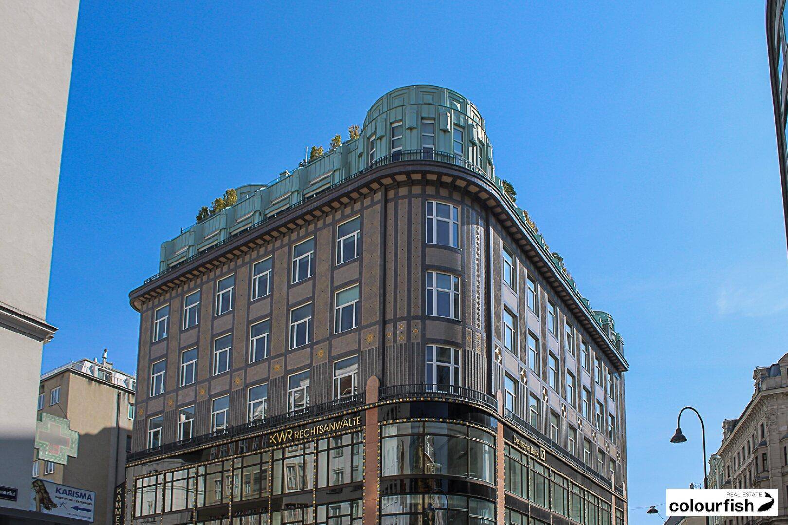 Historisches Gebäude mit markanter Architektur, runder Ecke und grünem Kupferdach unter blauem Himmel.