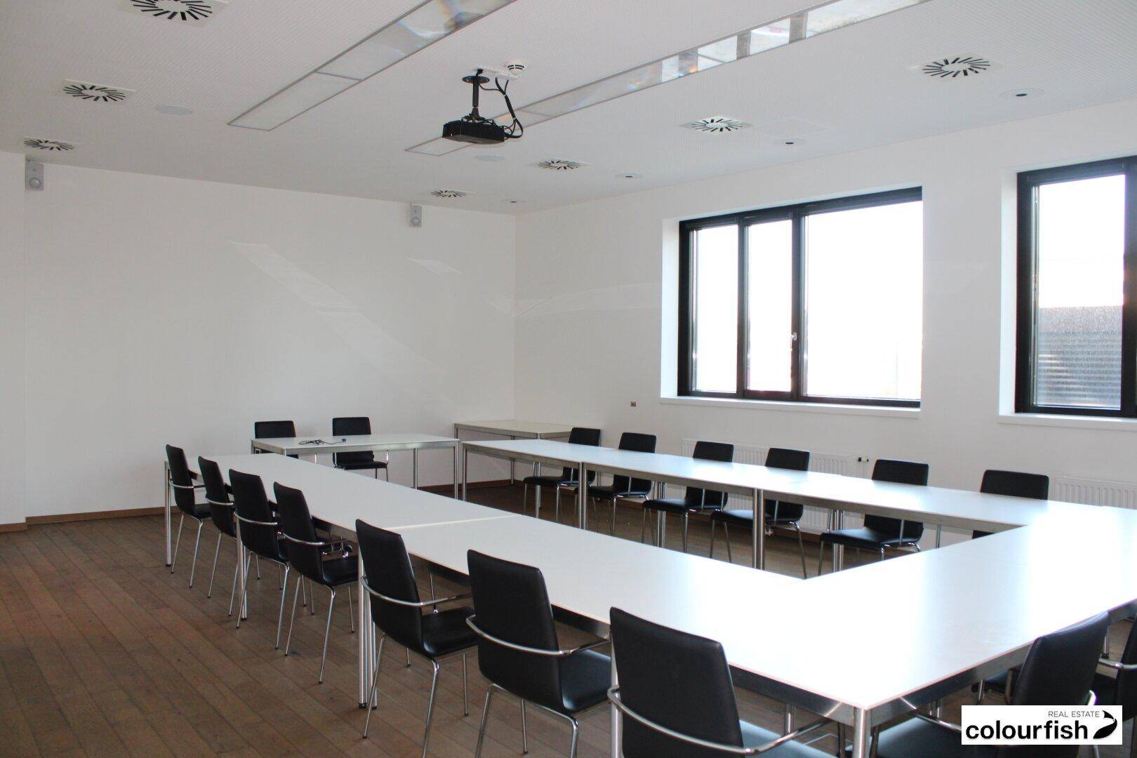 Geräumiger Konferenzraum aus einer anderen Perspektive, ausgestattet mit U-förmigen Tischen, Stühlen und Deckenlüftung.