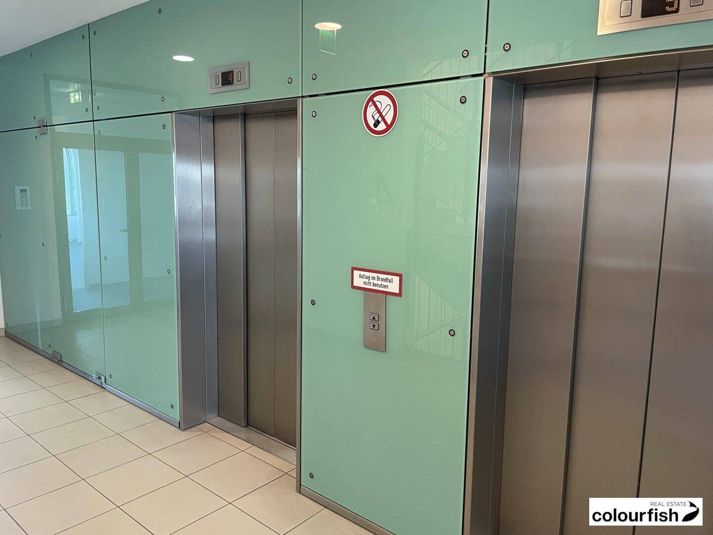 Moderner Aufzugsbereich mit glänzenden Metalltüren und hellgrünen Glaspaneelen in einem Bürogebäude.