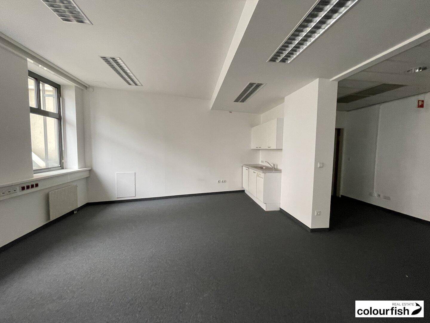 Büroraum mit integrierter Teeküche, Teppichboden und großen Fenstern für ausreichend Tageslicht.