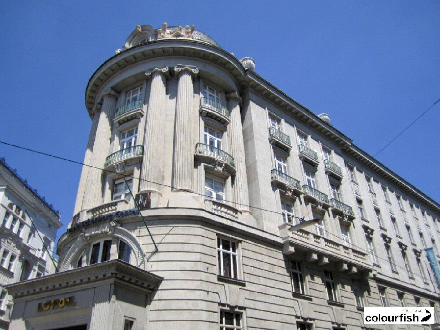 Prächtiges Eckgebäude mit klassischer Architektur, Säulen, kleinen Balkonen und blauem Himmel.