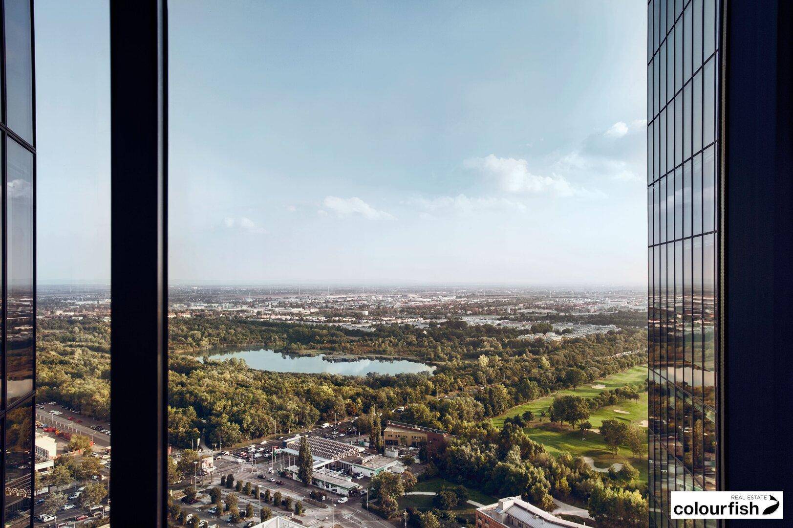 Panoramablick aus einem Hochhausfenster auf eine Stadtlandschaft mit See und Grünflächen.