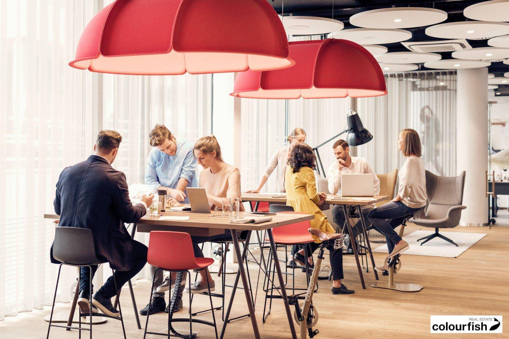Belebter Co-Working-Bereich mit großen roten Pendelleuchten, Holzboden und arbeitenden Personen.