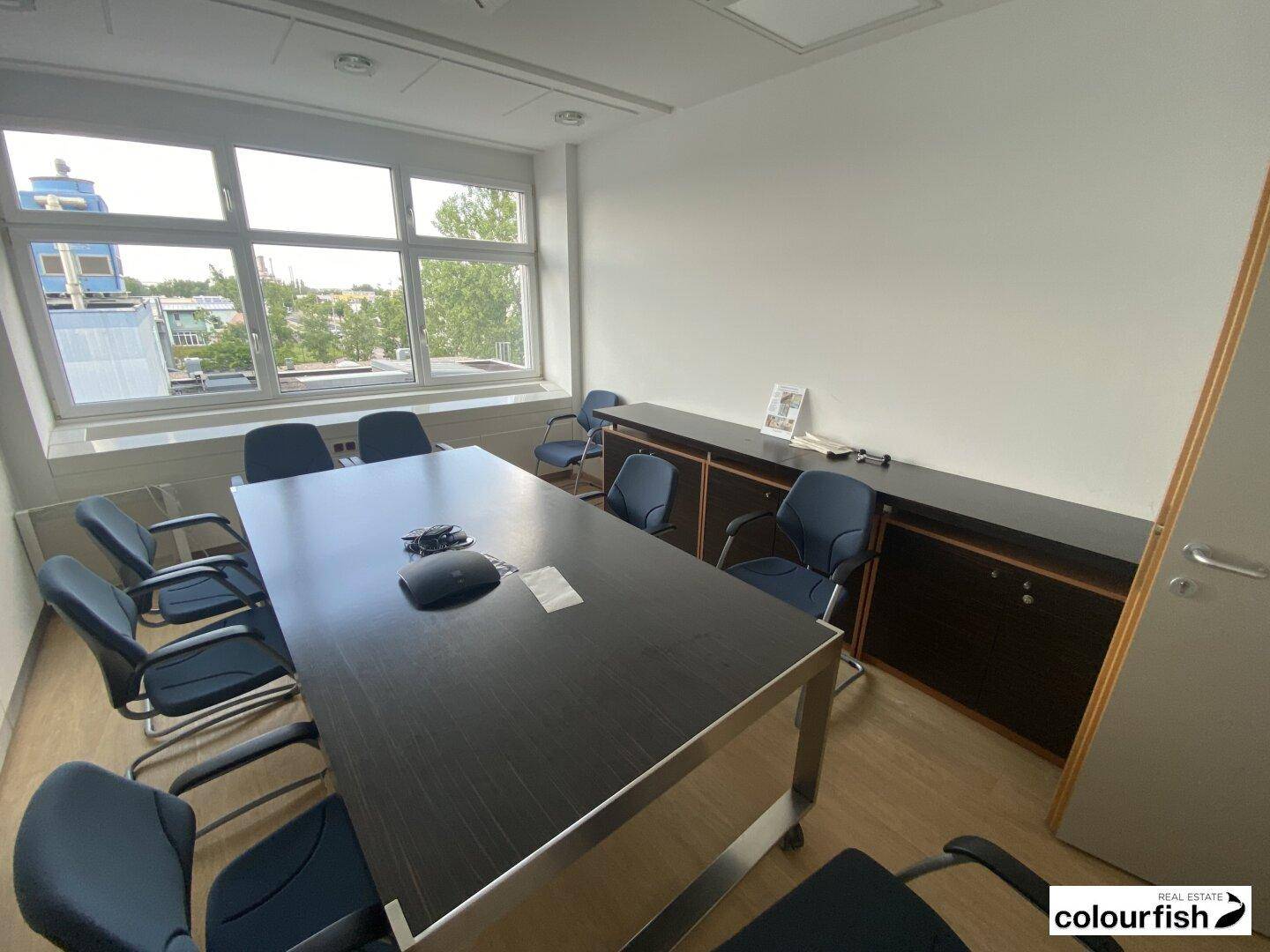 Konferenzraum mit großem Tisch, blauen Stühlen und Fenstern, die Tageslicht hereinlassen.