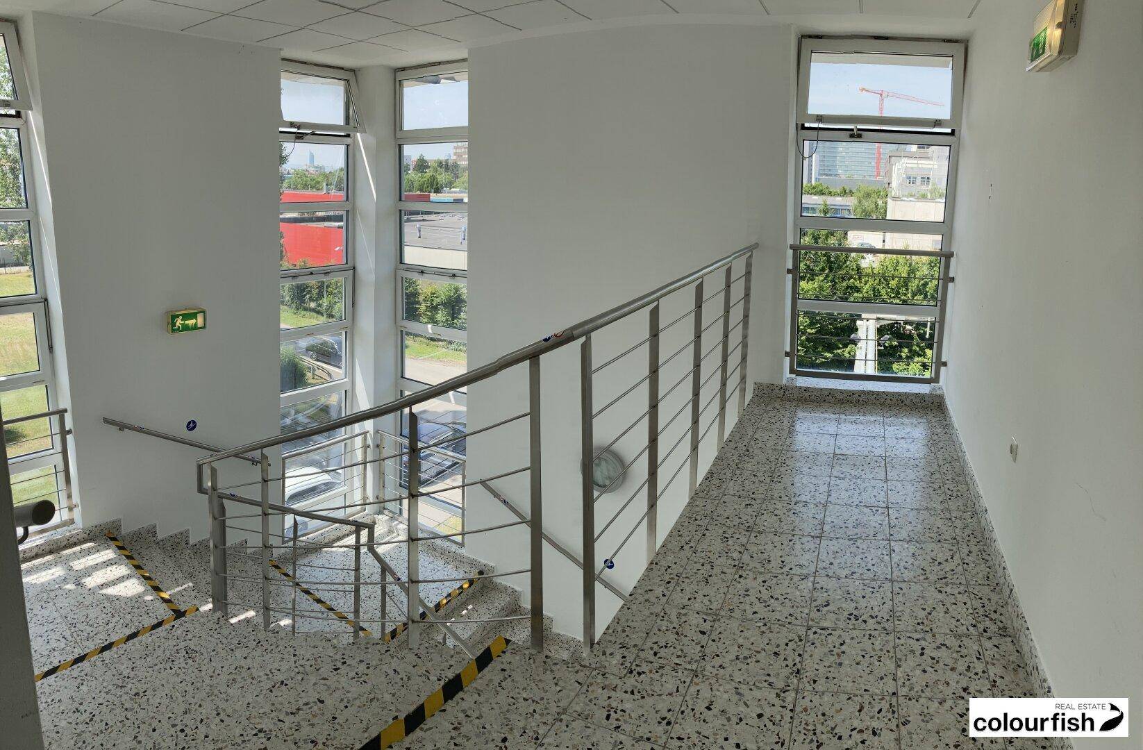 Helles Treppenhaus mit großen Fenstern, Metallgeländer und einem Terrazzo-ähnlichen Bodenbelag.