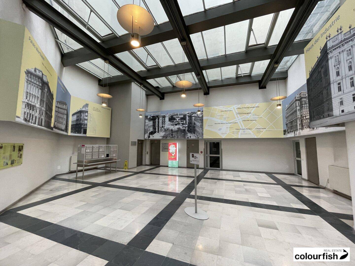 Geräumige, moderne Lobby mit Glasdach, Marmorboden und Informationsdisplays, viel Tageslicht.