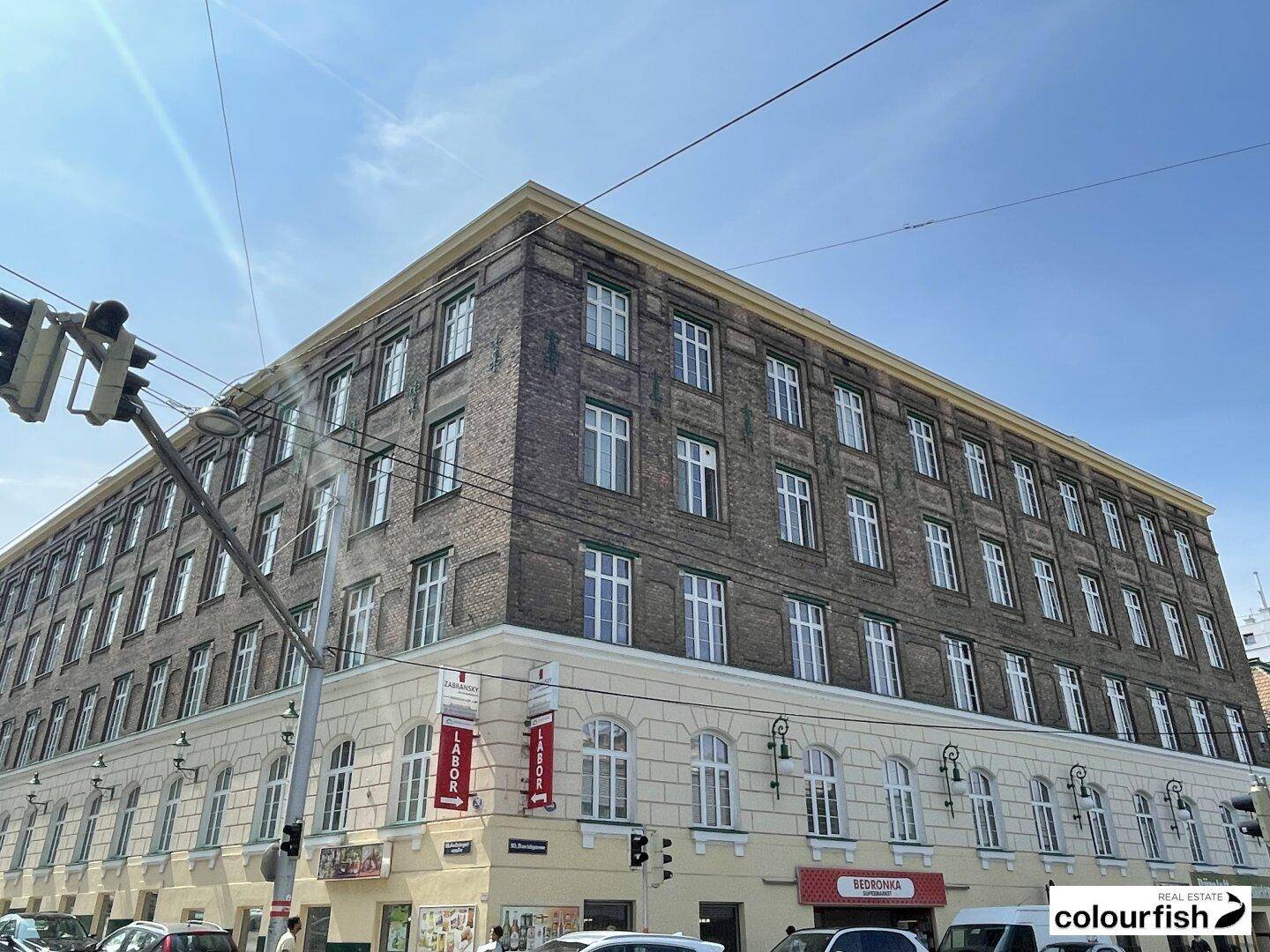 Historisches Gebäude mit markanter Fassade, großen Fenstern und Geschäften im Erdgeschoss an einer Straße.