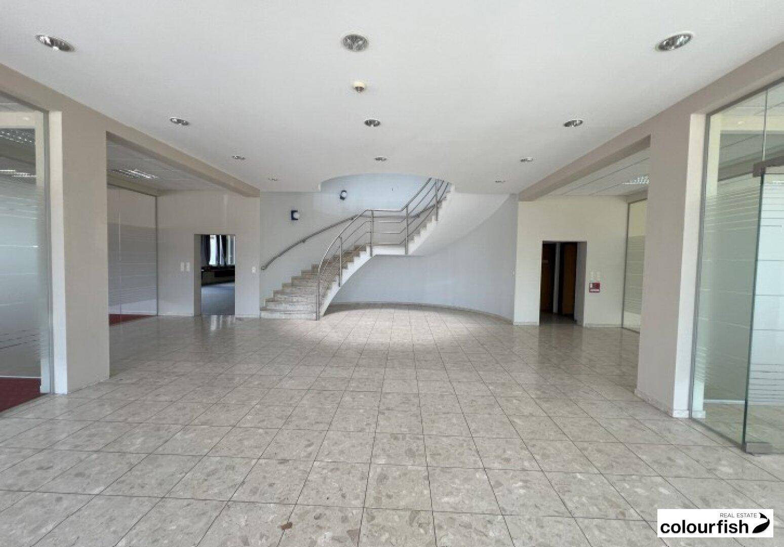 Heller Eingangsbereich mit Fliesenboden, geschwungener Treppe und Glaswänden zu angrenzenden Büros.