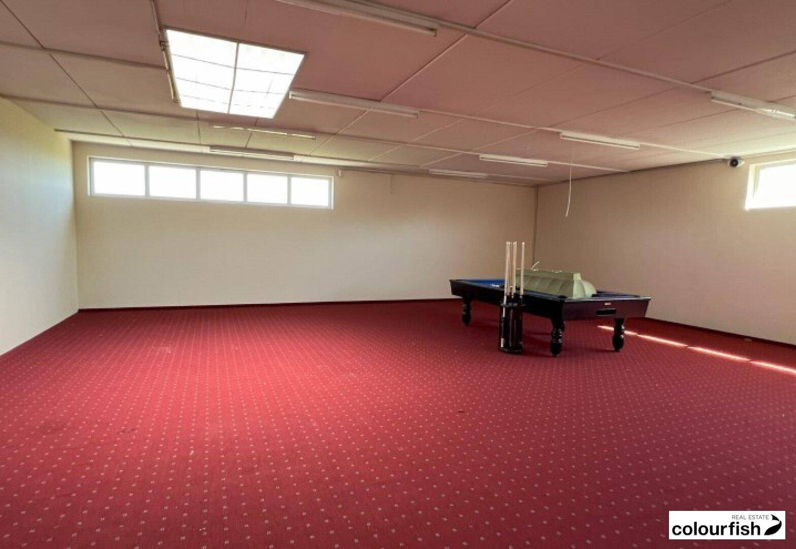 Großer Raum mit rotem Teppichboden, Billardtisch und kleinen Fenstern nahe der Decke.
