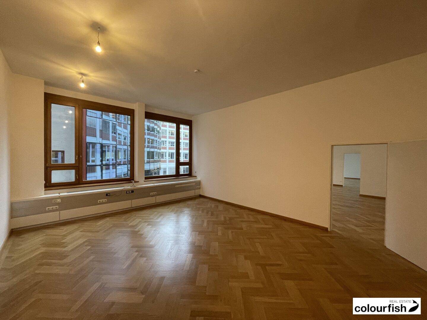 Geräumiger, leerer Raum mit Fischgrätparkett und großen Fenstern, die viel Tageslicht hereinlassen.
