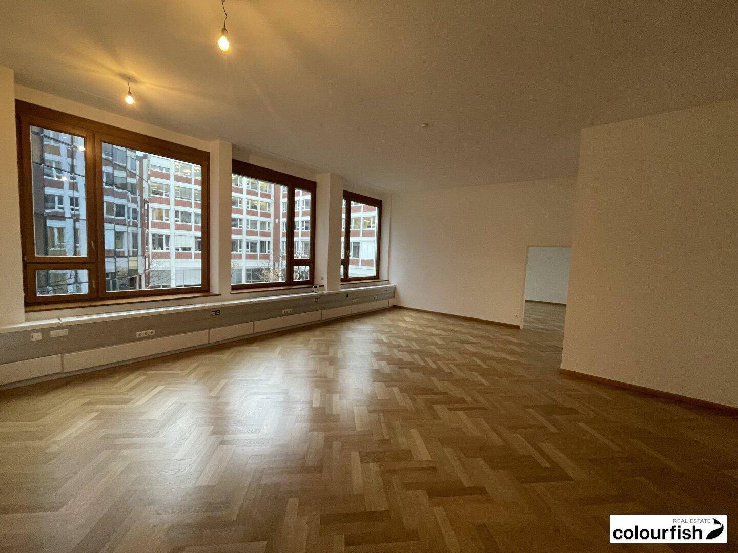 Heller, unmöblierter Raum mit klassischem Fischgrätparkett und breiten Fenstern mit Stadtblick.