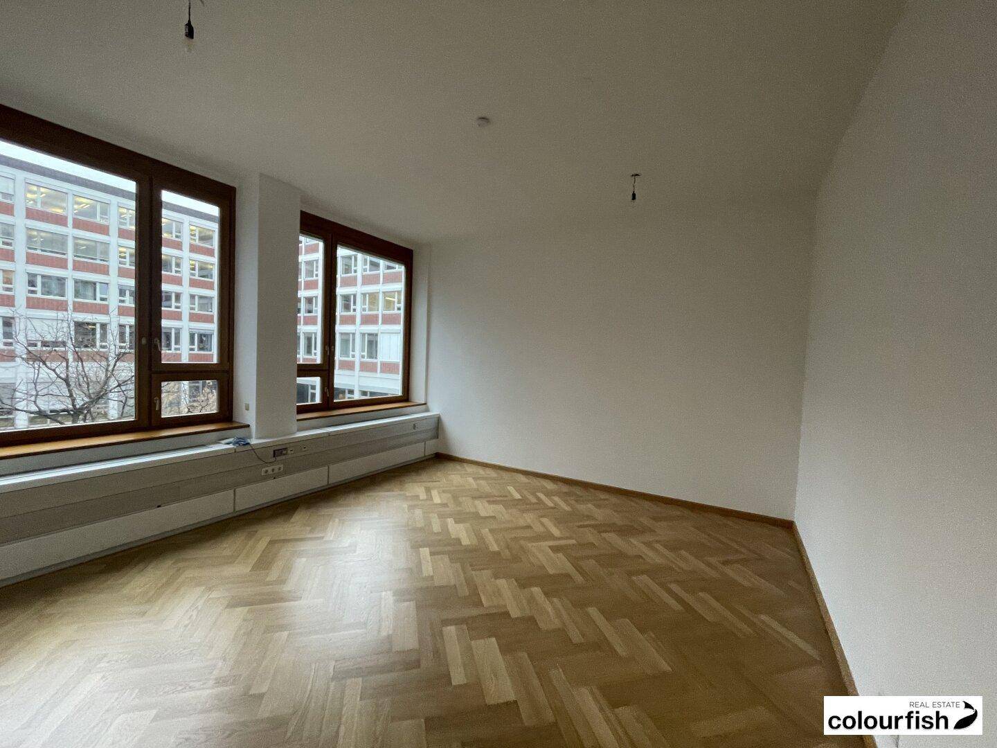 Lichtdurchfluteter Raum mit elegantem Fischgrätparkett und großen Fenstern, die eine städtische Aussicht bieten.