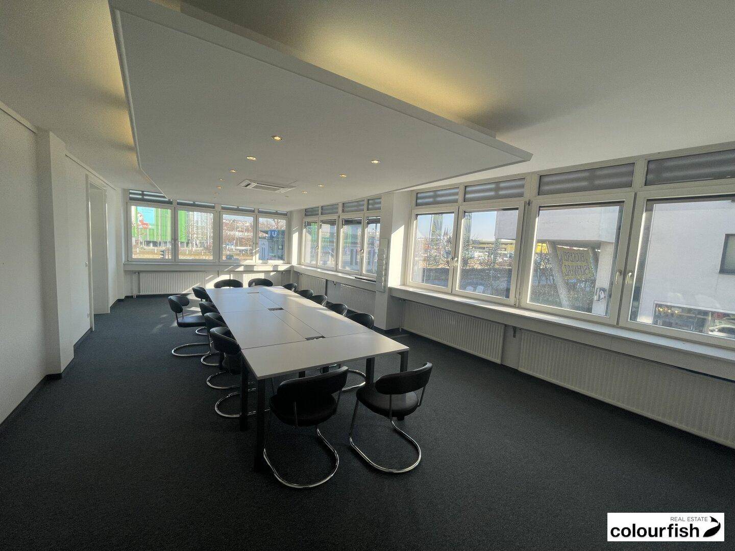 Geräumiger Konferenzraum mit großem Tisch, vielen Fenstern und Teppichboden.