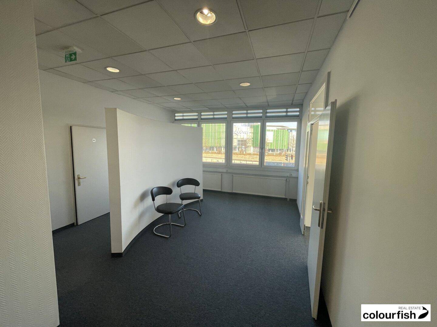 Offener Bürobereich mit zwei Stühlen, einer Trennwand und Fenstern.