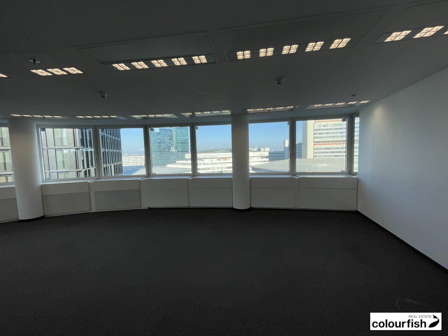 Leerer Büroraum mit Teppichboden und einer breiten Fensterfront, die einen Panoramablick auf die Stadt ermöglicht.