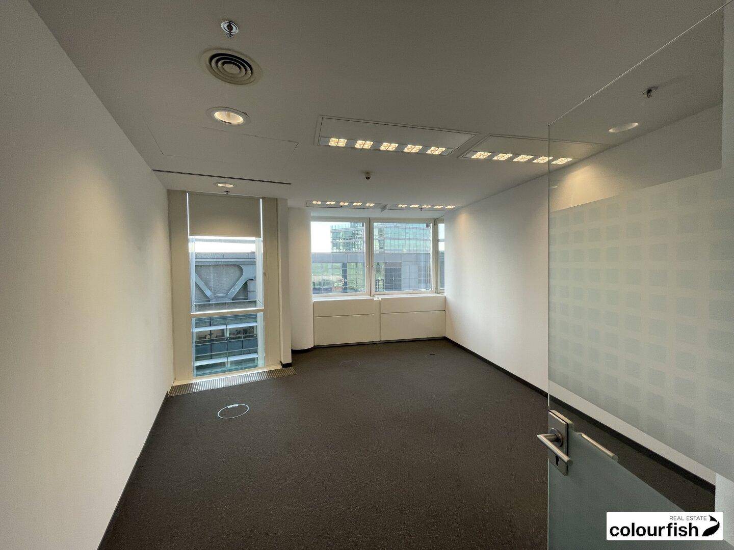 Helles Eckbüro mit Teppichboden, großen Fenstern und einer modernen Glaswand, die den Raum abgrenzt.