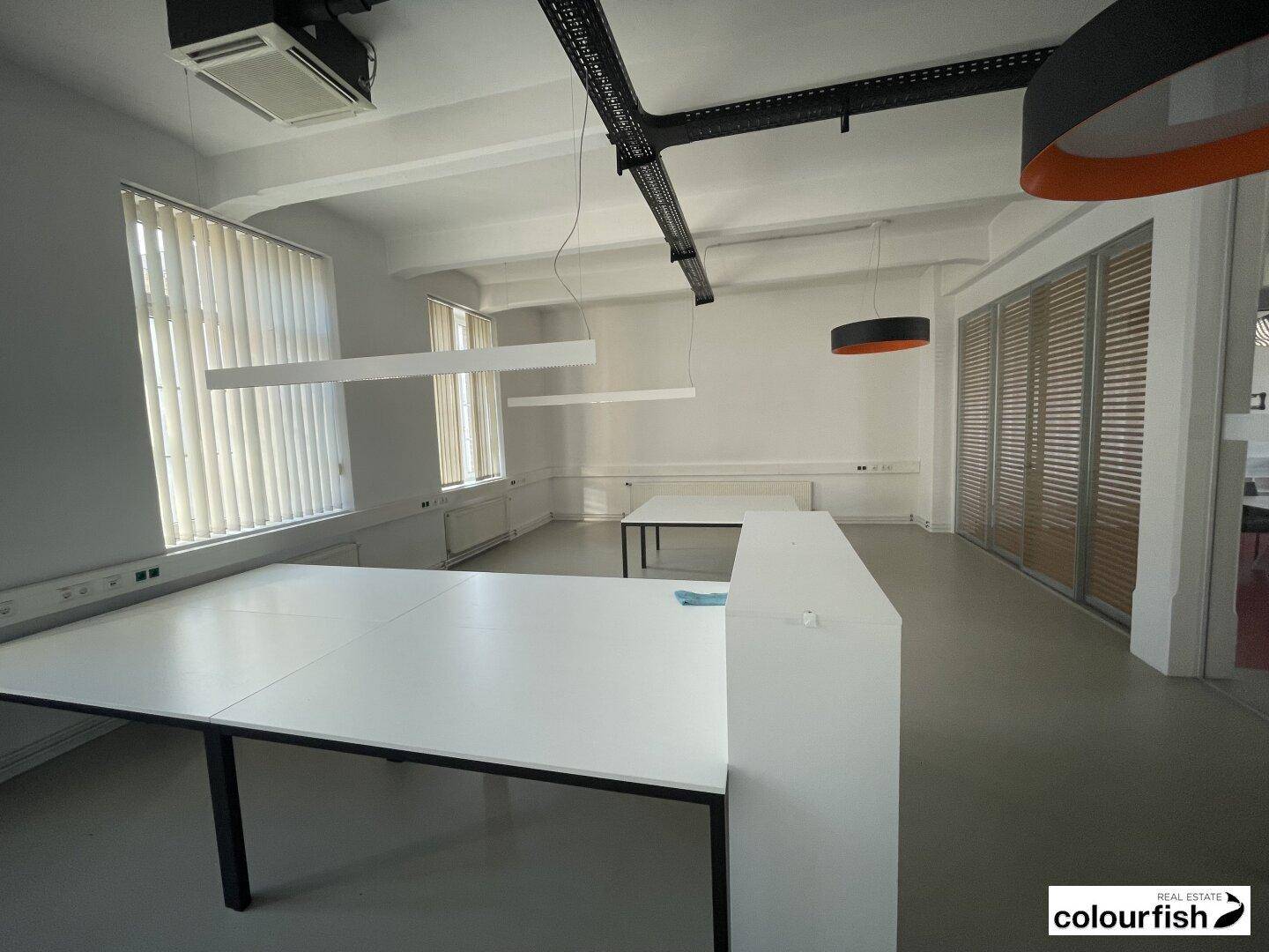 Helles Büro mit weißen Schreibtischen, vertikalen Jalousien an den großen Fenstern und moderner Beleuchtung.