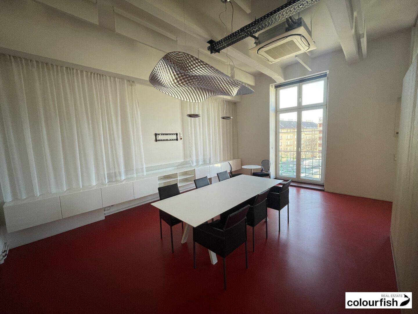 Moderner Konferenzraum mit rotem Boden, großem Tisch, Designerleuchte und bodentiefen Fenstern.