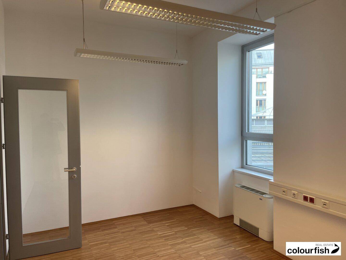 Leerer Büroraum mit Holzboden, weißen Wänden und einem großen Fenster, das viel Licht hereinlässt.