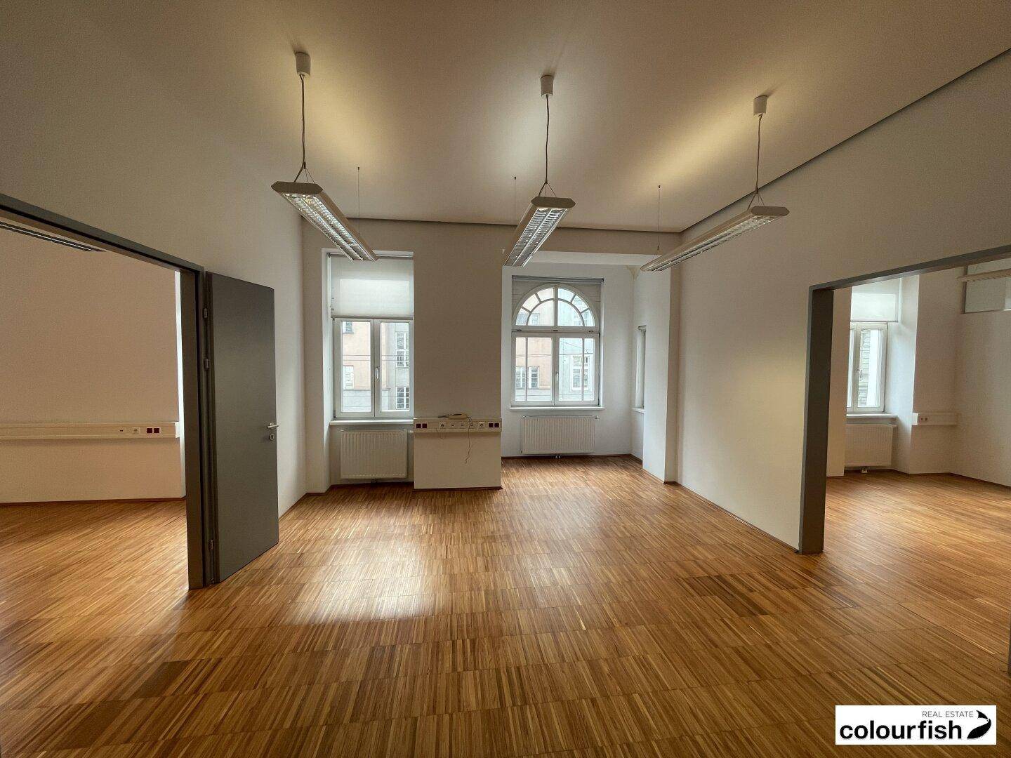 Heller Büroraum mit Holzboden und hohen Fenstern, darunter ein markantes Rundbogenfenster.