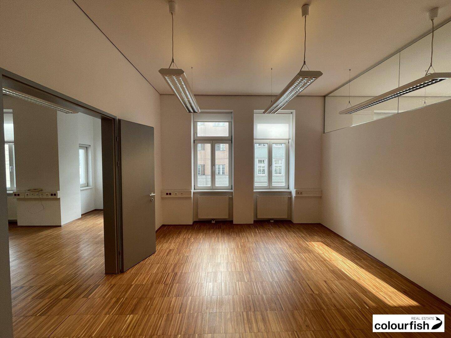 Leerer Büroraum mit Holzboden, weißen Wänden und zwei großen Fenstern, die den Raum erhellen.