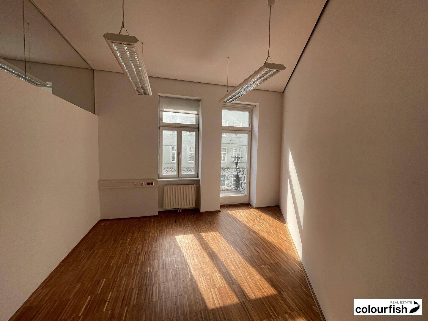 Heller Büroraum mit Holzboden und Fenstern, die zu einem kleinen französischen Balkon führen.