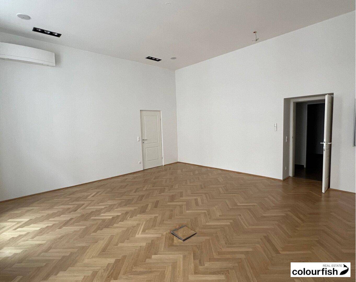 Großes, leeres Büro mit hellem Fischgrätparkett, weißen Wänden und einer Klimaanlage an der Decke.