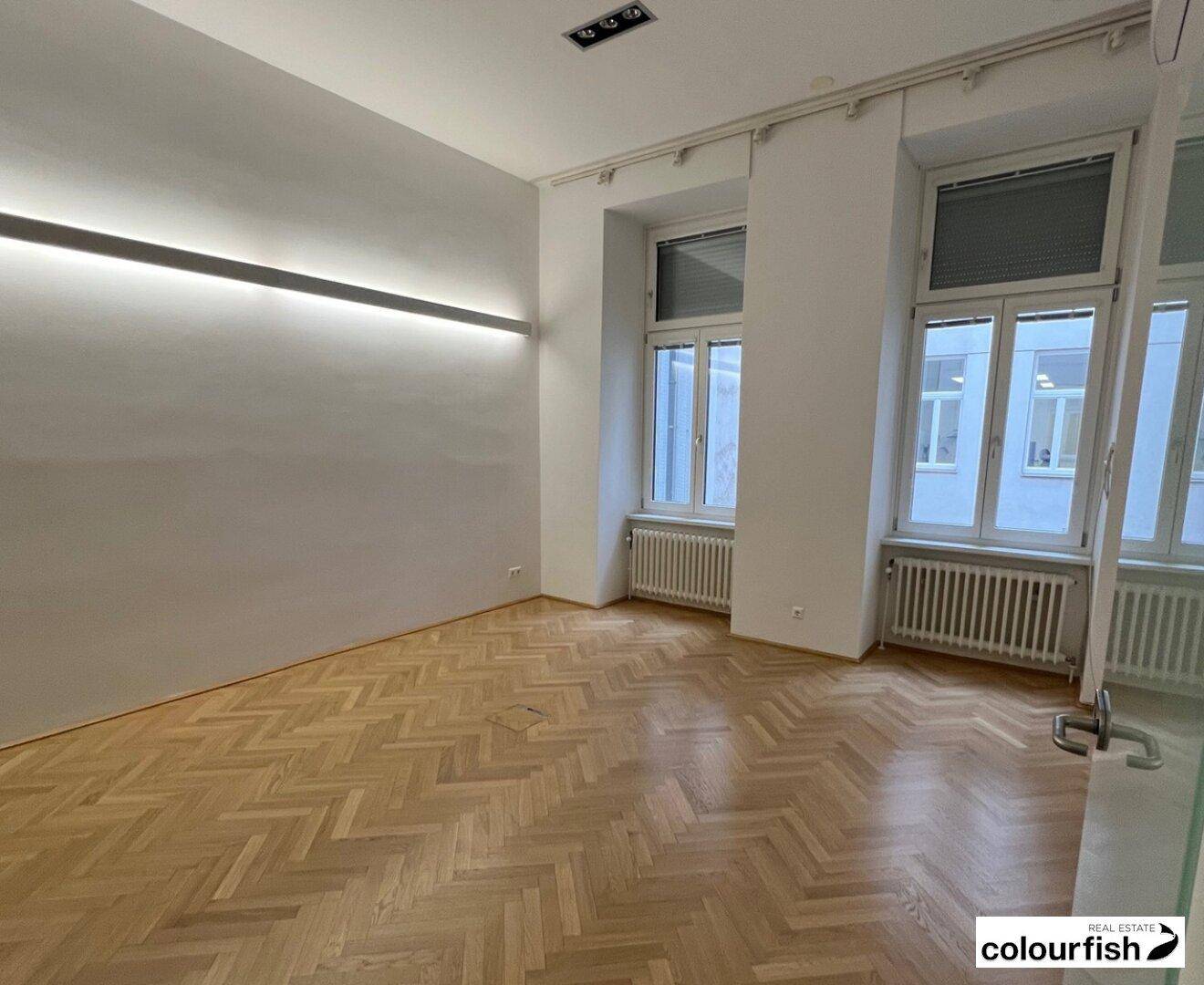Helles Büro mit Fischgrätparkett, zwei großen Fenstern und einer modernen Wandbeleuchtung.